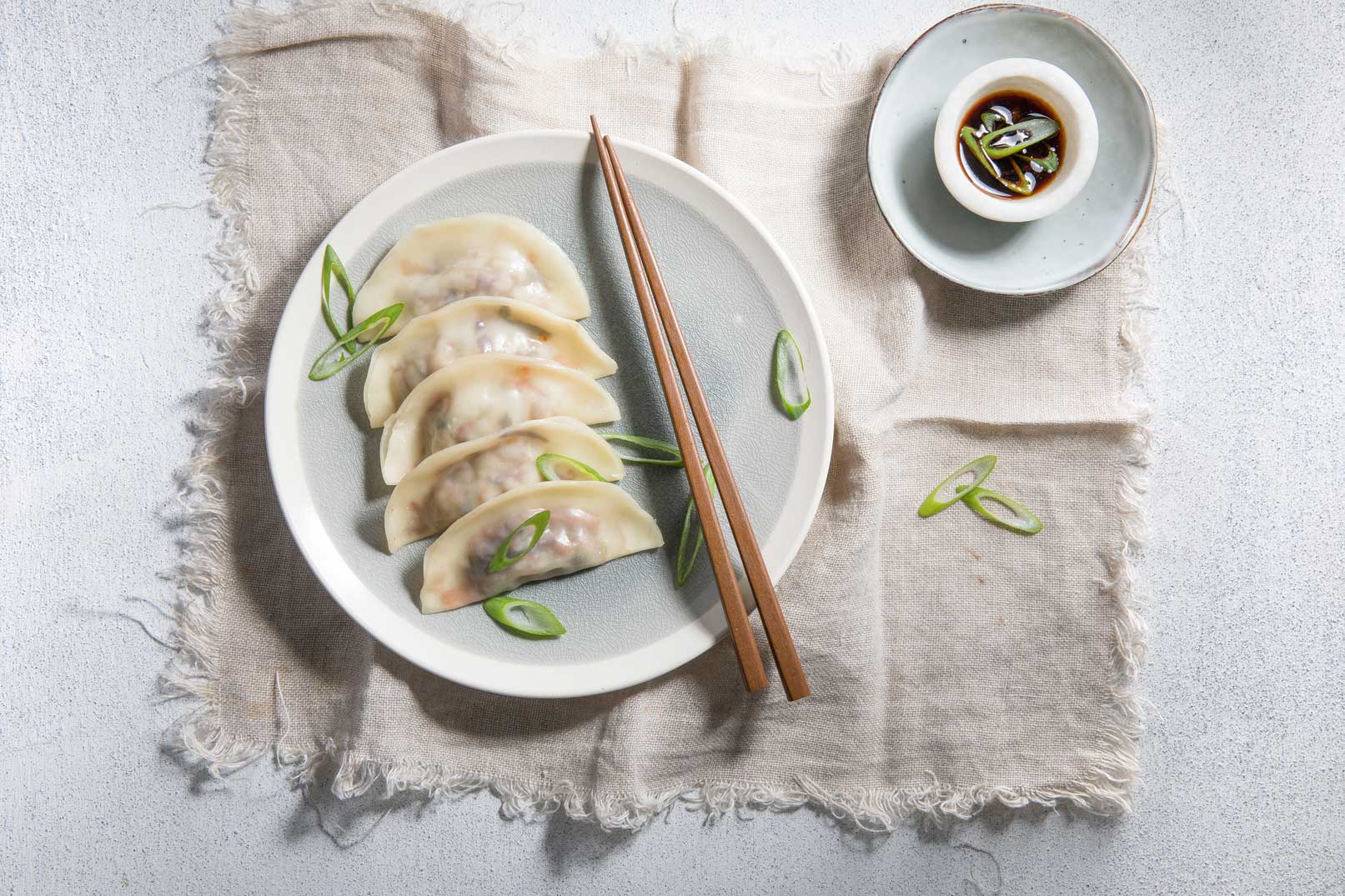 Five vegetable dumplings with sliced spring onion and wooden chopsticks on a large round plate, served on a rustic cloth napkin with a side dish of dipping sauce.