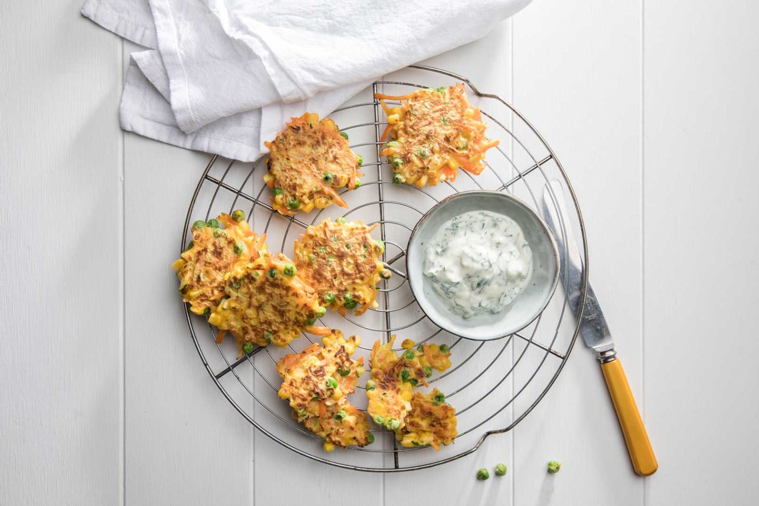 Vegetable fritters on a round wire cooling rack with tzatziki and a knife for spreading to serve.