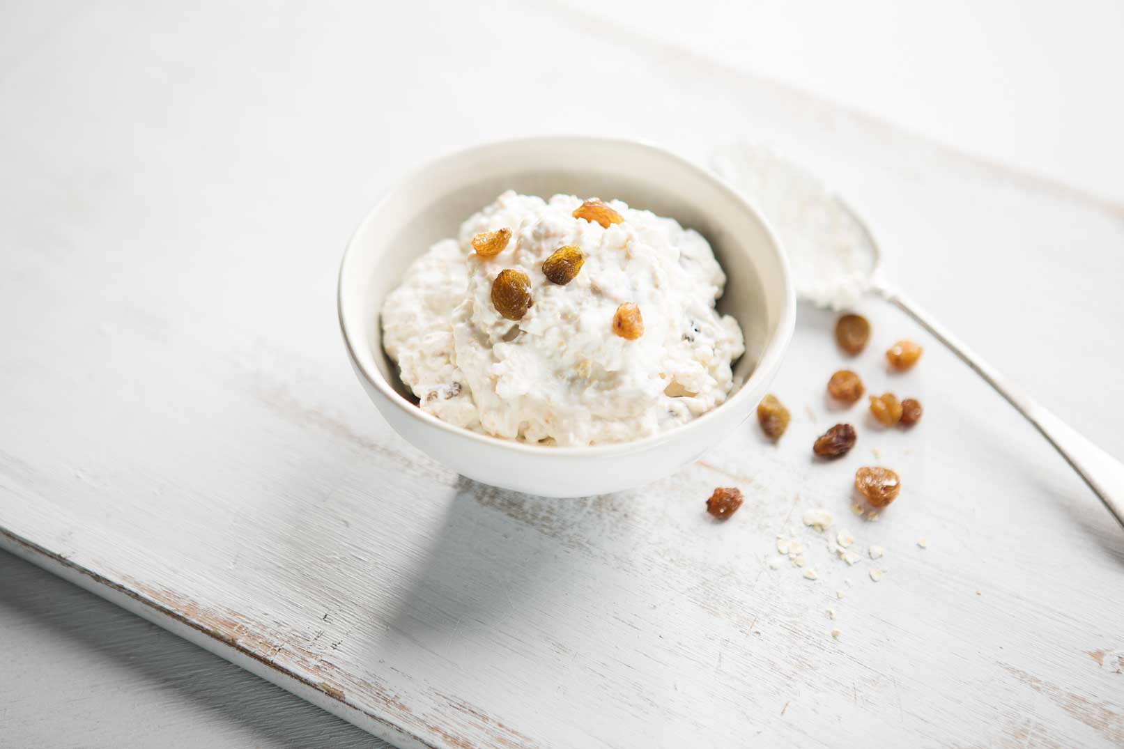 Mixed yoghurt and oats in a small white bowl topped with sultanas and a serving spoon on the side