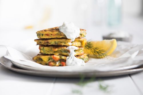 Image of stacked zucchini and corn fritters topped with tzatziki on a large plate with a side of dill and lemon wedges