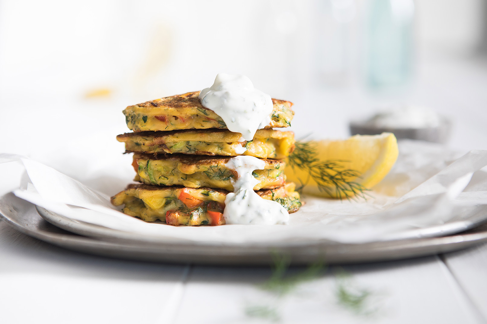 Image of stacked zucchini and corn fritters topped with tzatziki on a large plate with a side of dill and lemon wedges