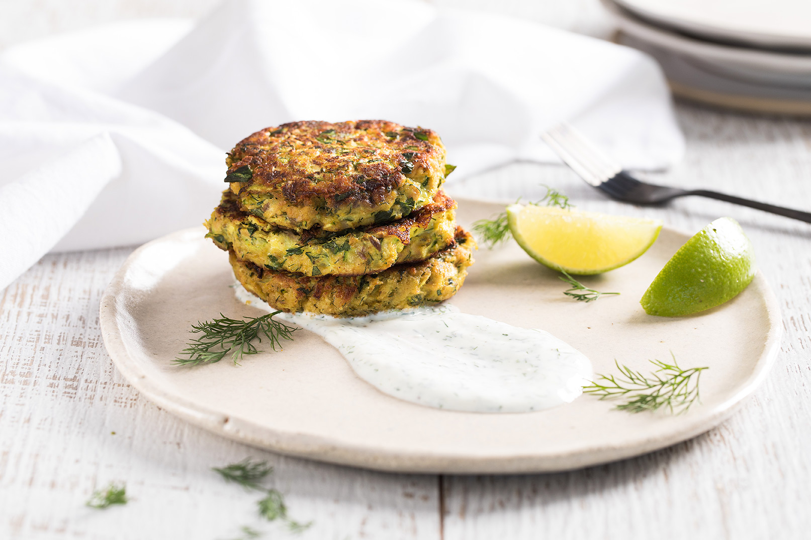 Image of stacked zucchini tumeric and salmon patties with a side of tzatziki and lemon wedges on a large stone plate. 