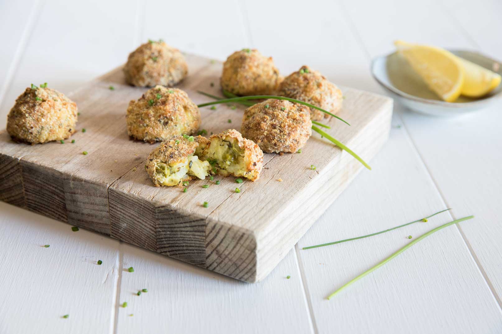 Cooked zucchini and cheese arancini balls served on a thick wooden cutting board with chives and a small dish of lemon wedges in the background for serving. 