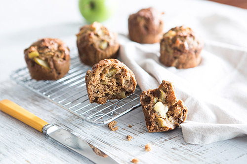 Image of baked apple and date muffins on a cooling rack with one open muffin with margarine spread and a knife.