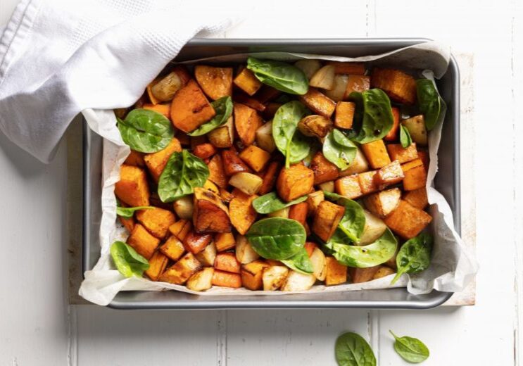 Roasted vegetables in a metal baking dish lined with baking paper, garnished with fresh spinach leaves