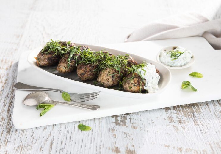 Image of five baked beef koftas on a long serving dish topped with Tzatziki and herbs with serving utensils on the side 