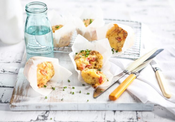 Image of baked easy savoury muffins in baking paper on a white cutting board and cooling rack with knives and a glass jug of water on the side. 