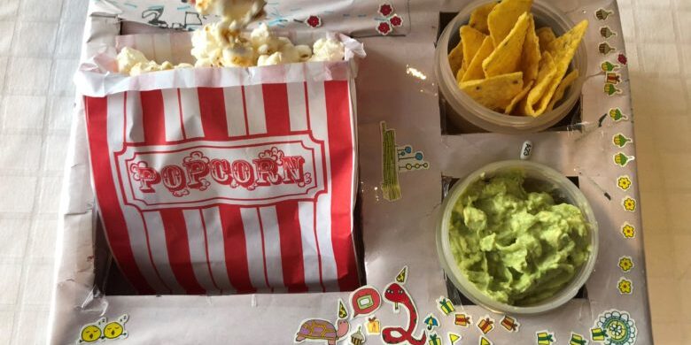 A cardboard box with cut outs for a bag of popcorn,guacamole and corn chips