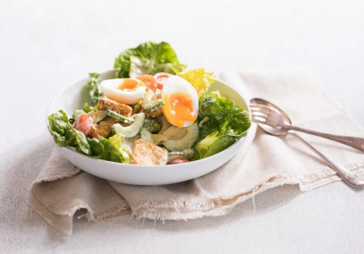 Vegetarian Caesar salad in a white bowl on a cloth napkin with a silver spoon and fork on the side.