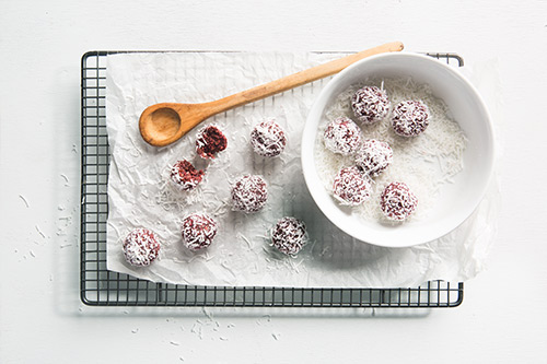 Image of six beetroot and spinach bliss balls in a bowl of coconut for rolling and another six balls on a baking paper on a cooling rack with a wooden spoon