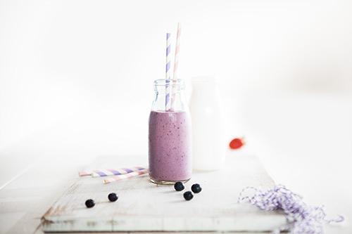 Image of a berrylicious smoothie in a glass milk jug with two straws, sitting on a white cutting board with blueberries, straws and string on the side as serving decorations