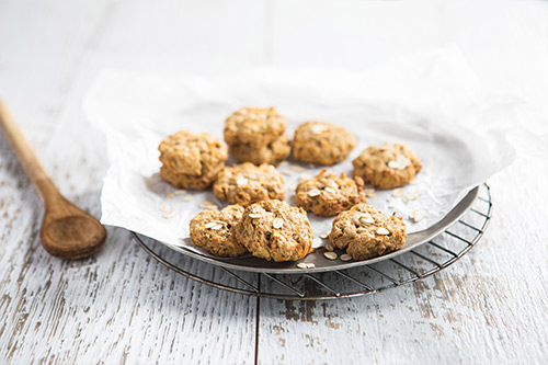 Image of 11 baked carrot cake oat biscuits sitting on baking paper on a round baking tray on a cooling rack with a wooden spoon on the side