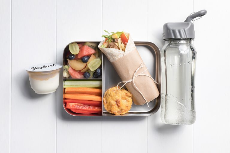 Image of a packed lunch box with a meatball and salad wrap, fruit salad, vegie sticks, easy savoury muffin and a yoghurt and bottle of water on the side