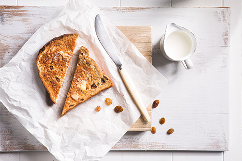 Image one slice of raisin bread sliced into two triangles served on baking baking and a cutting board with a knife and jug of milk on the side