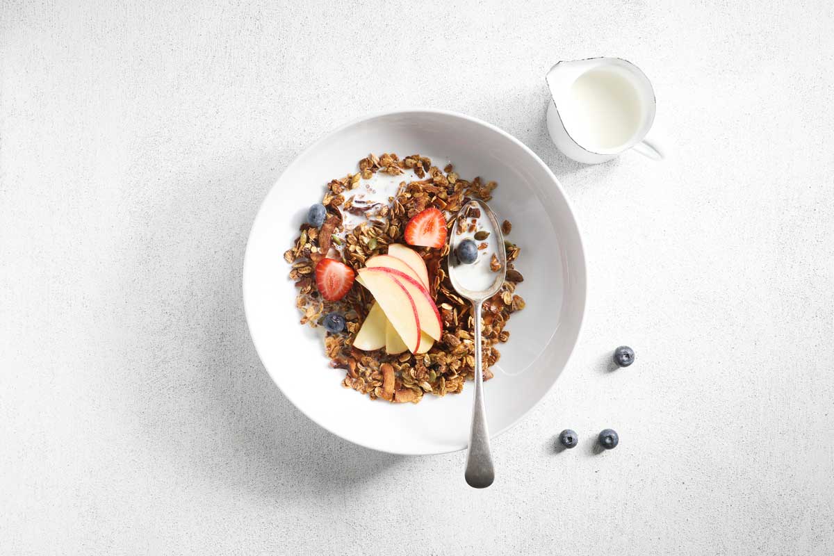Image of a white bowl of toasted muesli with a spoon in the bowl and a jug of milk on the side