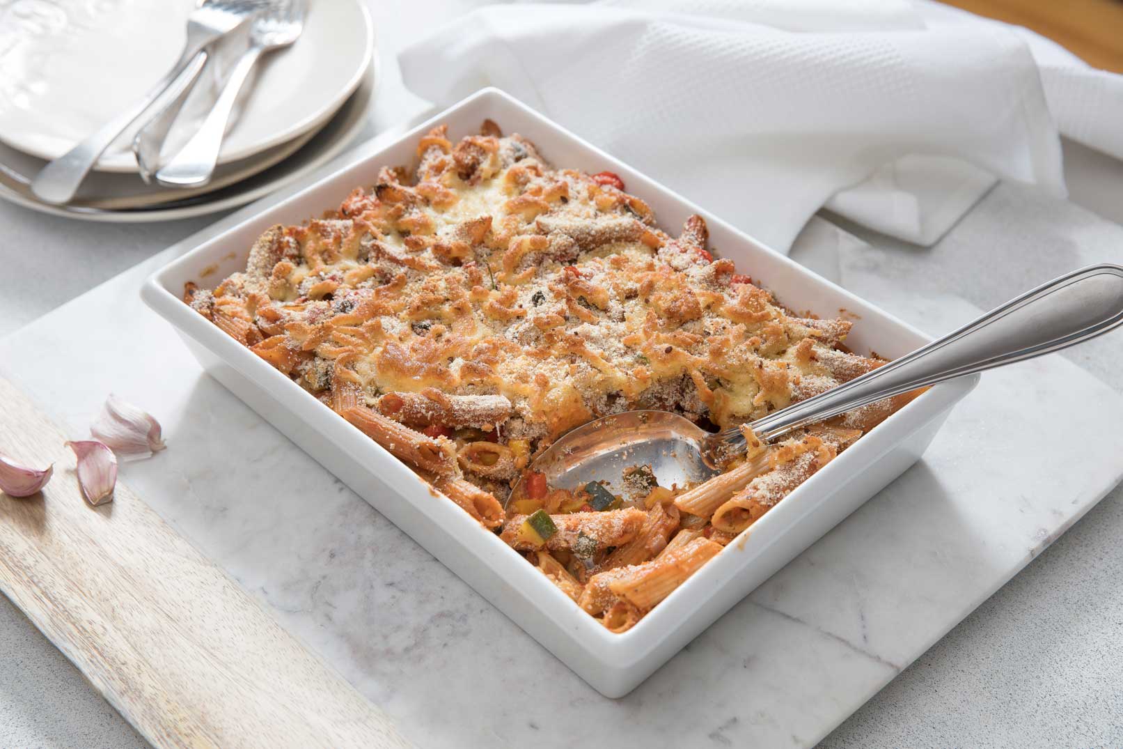 Vegetable pasta bake in a large white rectangle baking dish with a serving spoon and scoop taken out, served on a marble look cutting board with garlic cloves, three plates, cutlery and a white cloth napkin on the wide.